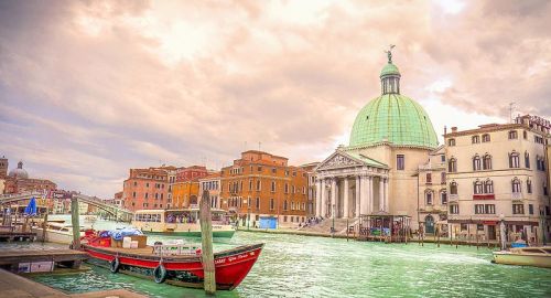 A scenic view of a canal in Venice, featuring a boat and a historic building with a green dome under a cloudy sky.