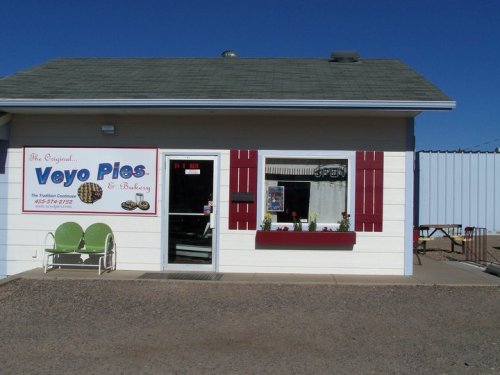 Exterior of Veyo Pies & Bakery, featuring a sign, open window, and green chairs on a sunny day.