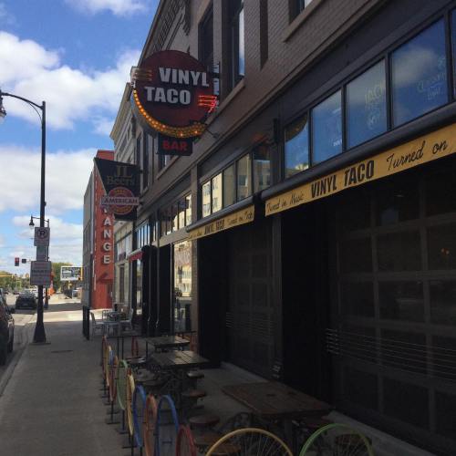Exterior view of Vinyl Taco bar with colorful outdoor seating and nearby signage for JL Beers and other establishments.