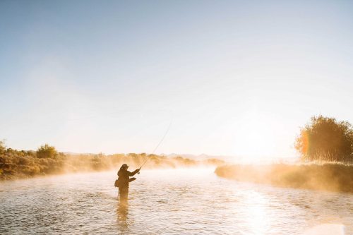A person fly fishing in a misty river at sunrise, surrounded by nature and soft golden light.