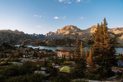 A serene mountain landscape at sunset, featuring a lake, rocky terrain, and a few tents among trees.
