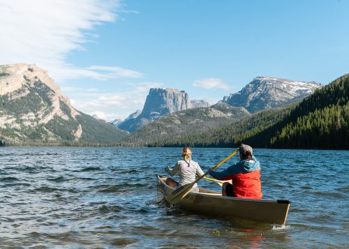 Two people paddle a canoe on a serene lake, surrounded by mountains and lush forests under a clear blue sky.
