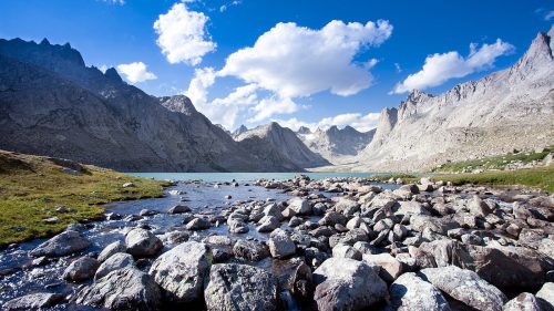 A serene mountain landscape with a clear lake, rocky shore, and dramatic peaks under a blue sky with fluffy clouds.