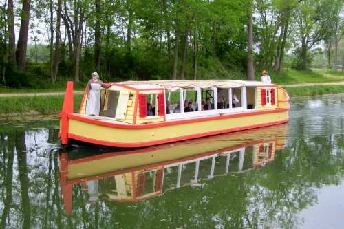 A colorful boat with passengers glides along a calm, tree-lined canal. Two people stand on the deck.