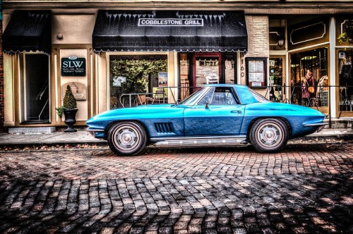 A vintage blue Corvette parked on a cobblestone street in front of a restaurant and shops.
