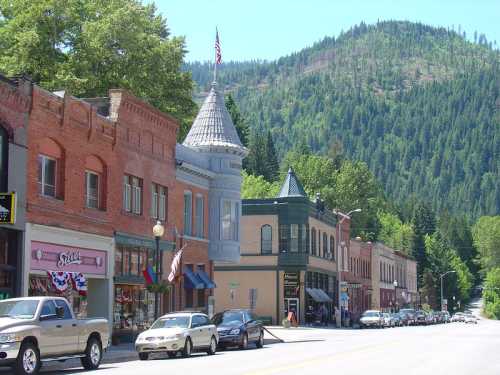 A charming small town street lined with historic buildings, shops, and trees, set against a backdrop of green mountains.