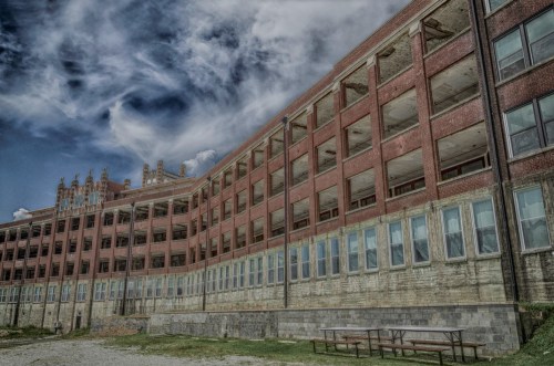 Abandoned brick building with large windows under a cloudy sky, featuring a weathered facade and grassy area in front.