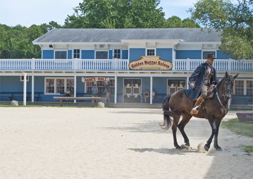 A cowboy rides a horse in front of a blue saloon with a sandy area and people sitting on the porch.