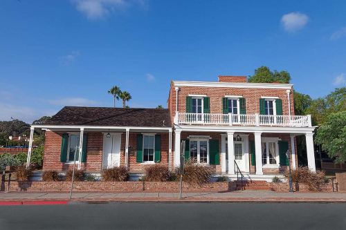 Historic brick building with green shutters, a porch, and palm trees under a blue sky.