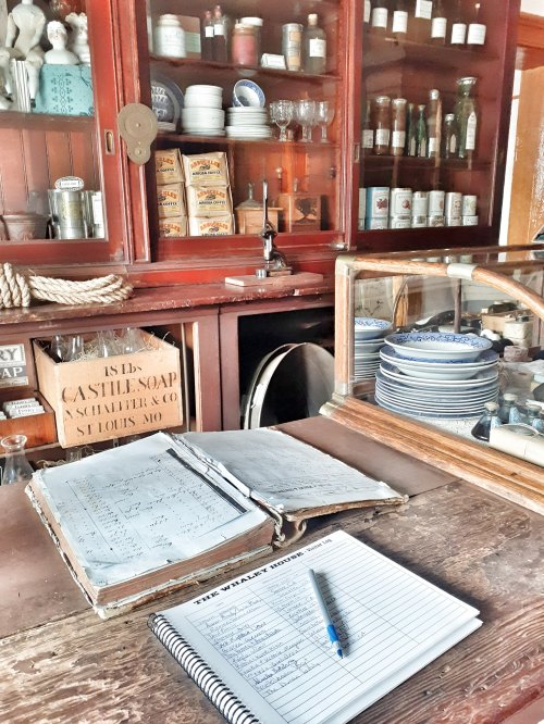 A vintage store interior with shelves of jars and bottles, an open ledger, and a notebook on a wooden counter.
