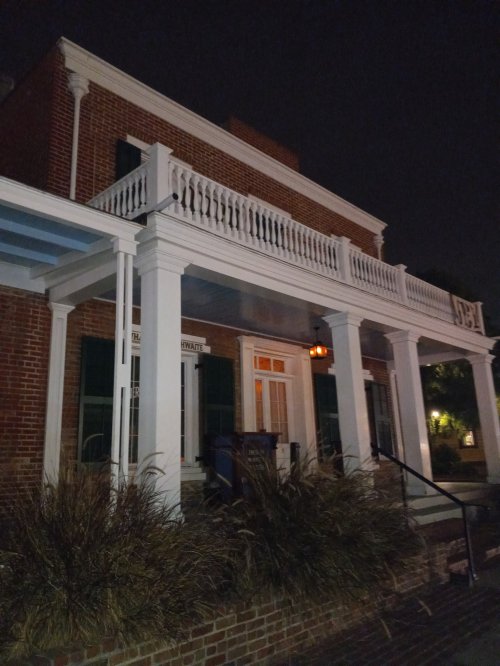 A brick building at night with a white porch, green shutters, and a warm light illuminating the entrance.