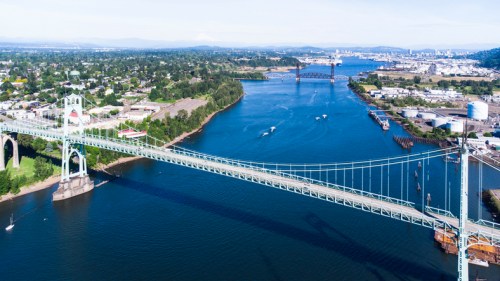 Aerial view of a bridge spanning a river, surrounded by greenery and urban areas, with boats navigating the water.