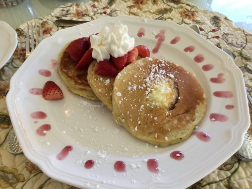 A plate of pancakes topped with strawberries, whipped cream, and powdered sugar, drizzled with fruit sauce.