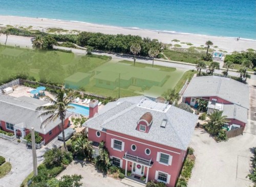 Aerial view of a pink beach house near the ocean, with palm trees and a pool in the foreground.