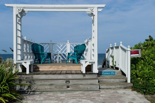A white gazebo with two teal chairs overlooks the ocean, surrounded by greenery and a sign nearby.