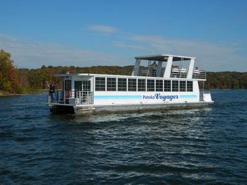 A boat named "Patoka Voyager" glides across a lake surrounded by autumn-colored trees under a clear blue sky.