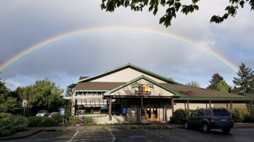 A building with a green roof under a vibrant rainbow, surrounded by trees and a cloudy sky.