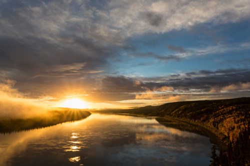 A serene landscape featuring a river reflecting a vibrant sunset, surrounded by trees and rolling hills under a cloudy sky.