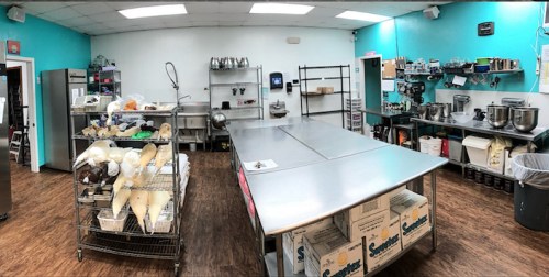 A bright kitchen with stainless steel tables, shelves of ingredients, and various kitchen equipment against turquoise walls.
