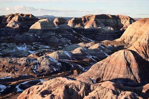 A panoramic view of colorful, layered rock formations and hills under a clear sky, with some snow patches visible.