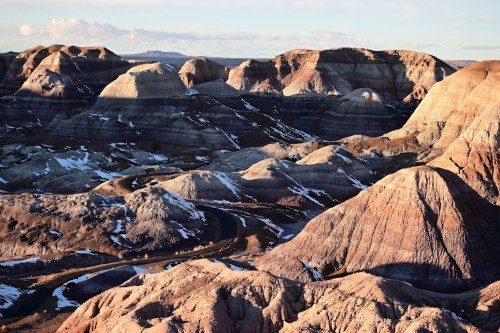 A panoramic view of colorful, layered rock formations and hills under a clear sky, with some snow patches visible.