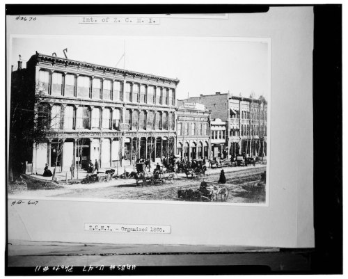 Historic black-and-white photo of a street scene featuring buildings, horse-drawn carriages, and people from the 1800s.
