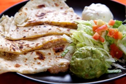 Three stuffed tortillas on a black plate, accompanied by salad, guacamole, and a dollop of sour cream.