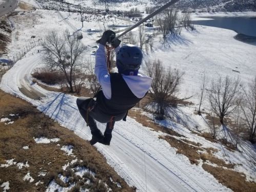 A person zip-lining over a snowy landscape with trees and a lake in the background.