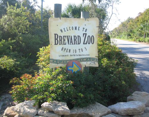 Sign welcoming visitors to Brevard Zoo, with operating hours and a directional arrow, surrounded by greenery.