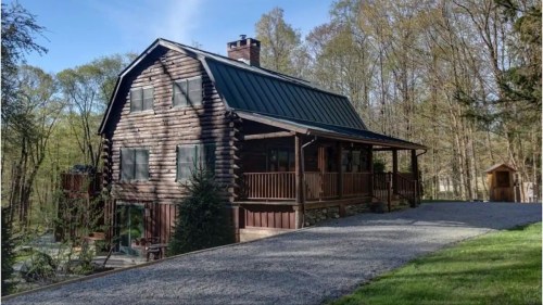 A rustic log cabin with a green metal roof, surrounded by trees and a gravel driveway.