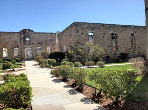 Ruins of a stone building surrounded by well-maintained gardens and pathways under a clear blue sky.