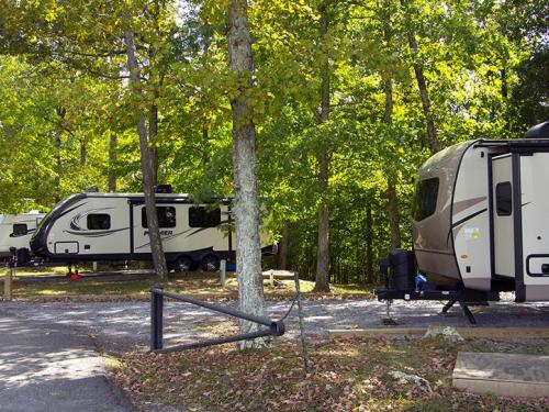 Two RVs parked in a wooded area, surrounded by trees and gravel paths, with sunlight filtering through the leaves.