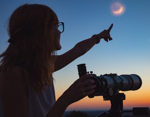 A silhouette of a woman pointing at the moon while holding a telescope against a sunset sky.