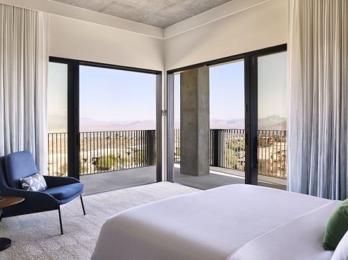 Modern bedroom with large windows offering a scenic view, featuring a bed, chair, and minimalist decor.