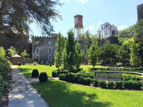 A lush green garden with neatly trimmed hedges, a stone building, and a water tower in the background under a clear blue sky.