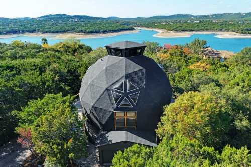 Aerial view of a unique dome-shaped house surrounded by trees, overlooking a serene lake and hills in the background.