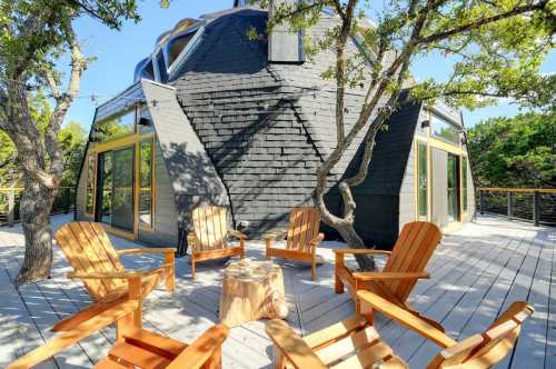 A modern, angular black house surrounded by trees, with wooden chairs arranged on a deck.