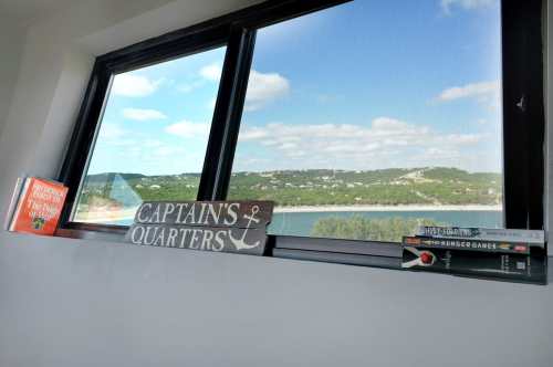 A window view overlooking a scenic landscape, with books and a sign reading "CAPTAIN'S QUARTERS" on the sill.