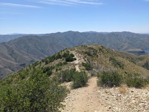 A scenic view of rolling hills and mountains under a clear blue sky, with a dirt path leading through greenery.