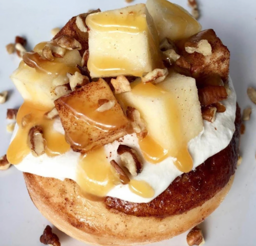 A cinnamon roll topped with cream, diced apples, pecans, and drizzled with caramel sauce.