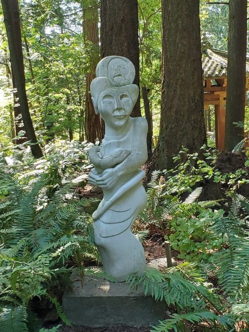 A tall, abstract stone sculpture of a figure holding a bird, surrounded by lush greenery and trees.
