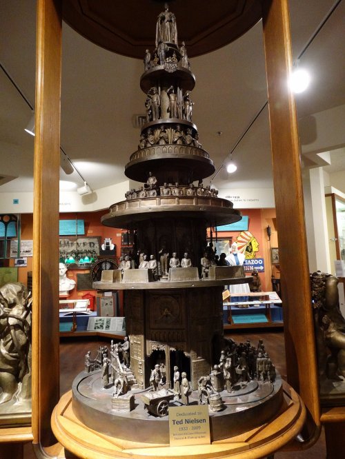 A multi-tiered sculpture featuring intricate figures, displayed in a museum with a plaque honoring Ted Nielsen.