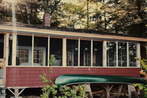 A cozy cabin with a screened porch, surrounded by trees, and a green canoe resting on a wooden support.