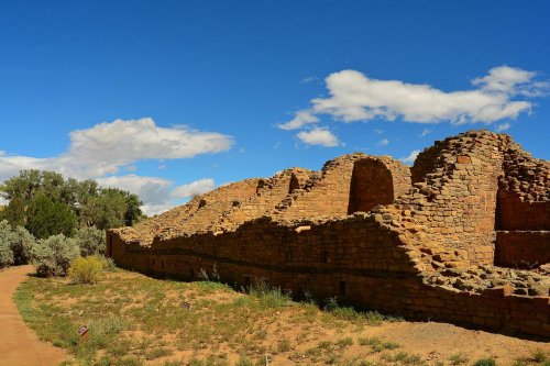 Ancient stone ruins under a blue sky with clouds, surrounded by desert vegetation and a winding path.