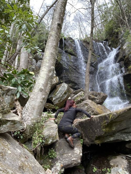 A person climbs over rocks near a waterfall surrounded by trees and greenery.