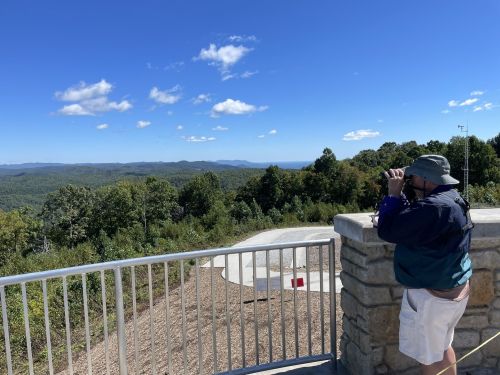 A person using binoculars to view a scenic landscape from a lookout point surrounded by trees and mountains.