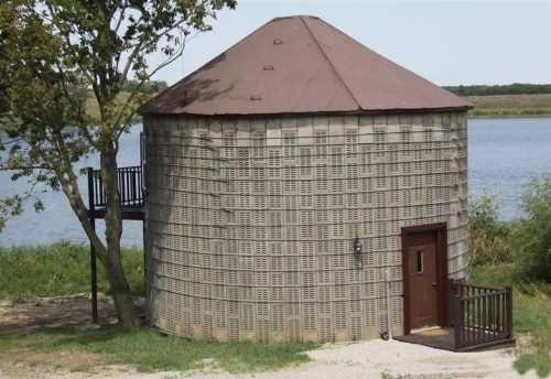 A round, silo-like structure by a river, featuring a brown roof and a wooden deck with stairs. Greenery surrounds it.