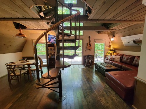 Cozy interior of a cabin featuring a spiral staircase, wooden accents, a seating area, and large windows with greenery outside.