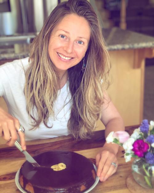 A smiling woman with long hair cuts a chocolate cake on a wooden table, surrounded by flowers.
