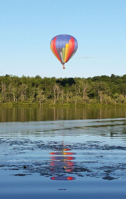 A colorful hot air balloon floats above a calm lake, reflecting on the water's surface with trees in the background.